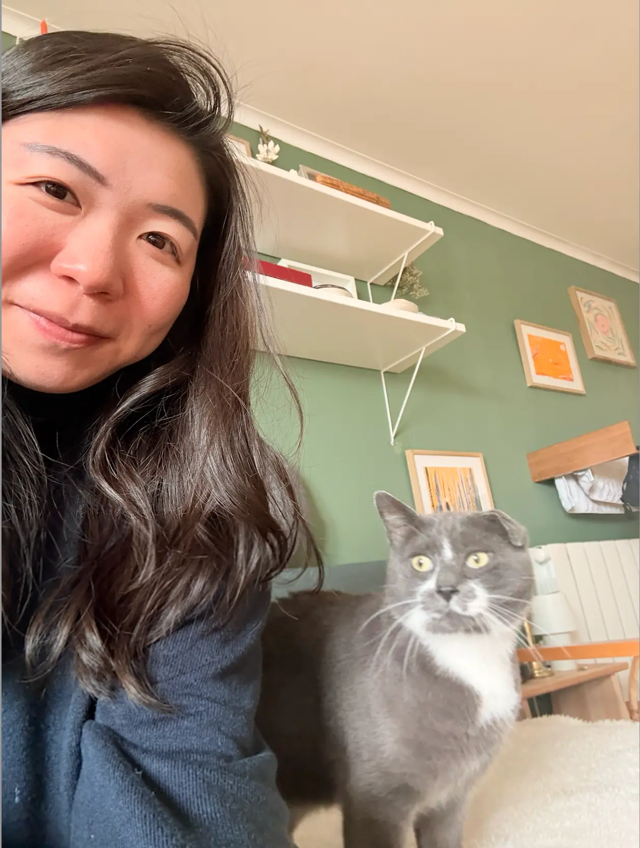 Chiu smiling at the camera with Sesame, a grey and white cat, standing beside her in a green-walled room